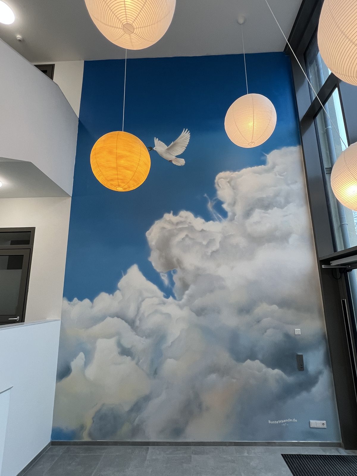 Foyer der Arche Tecklenburg mit Wandbild: Wolkenhimmel und Taube mit Olivenzweig im Schnabel, davor Papierlampen