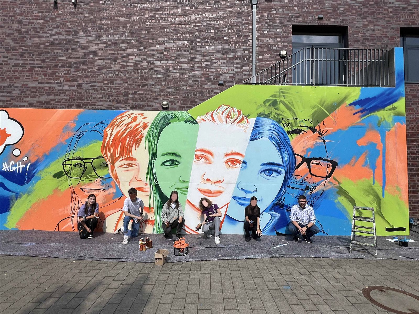 Schülerinnen und Schüler vor ihrem fertigen Graffiti-Workshop-Mural an der Harkenberg-Gesamtschule Hörstel: bunte Portraits im Pop-Art-Stil