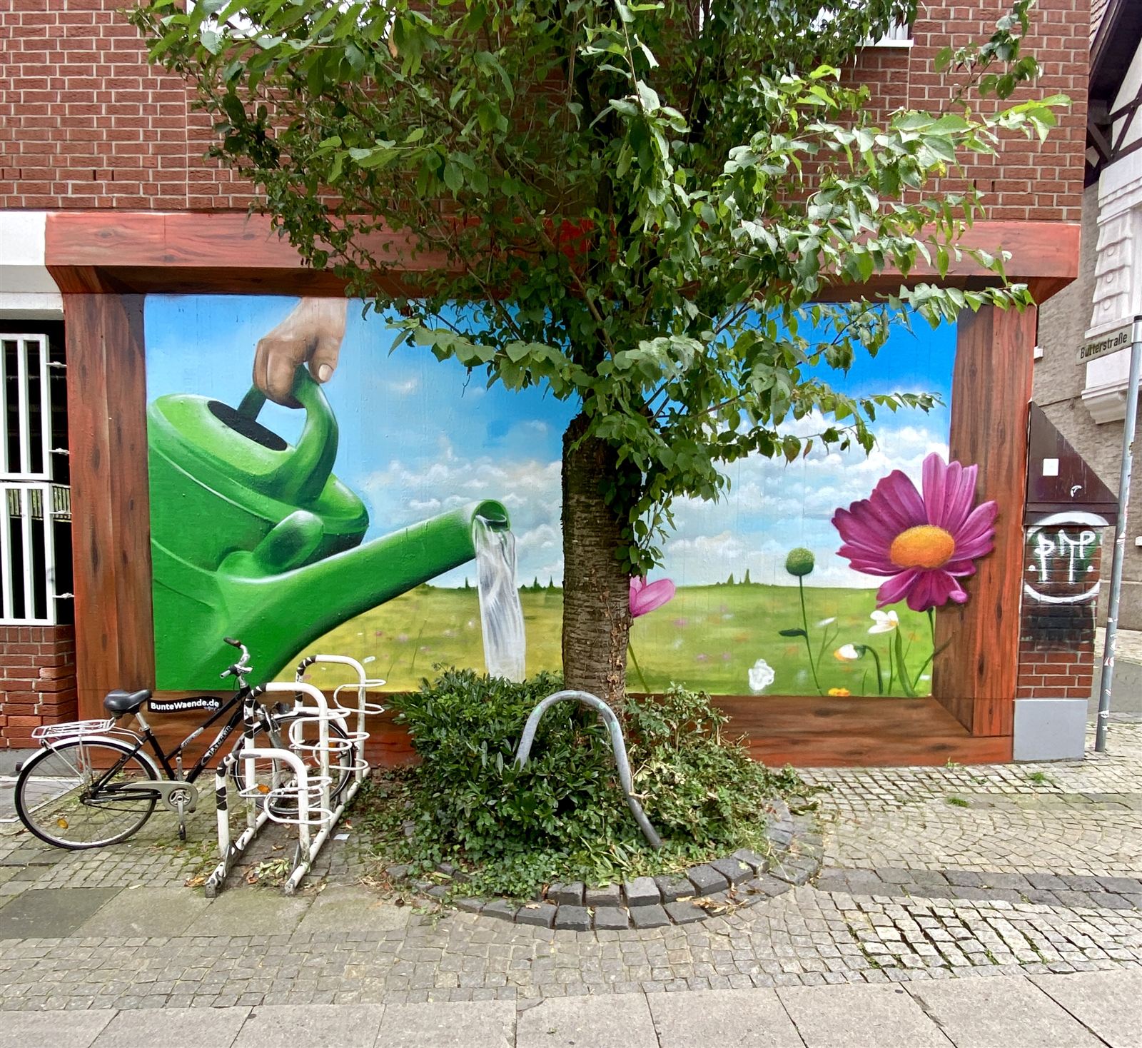 Mural an der Stadtkirche in Rheine: Gießkanne bewässert eine Sommerwiese, der Baum vor der Wand ist Teil des Bildes